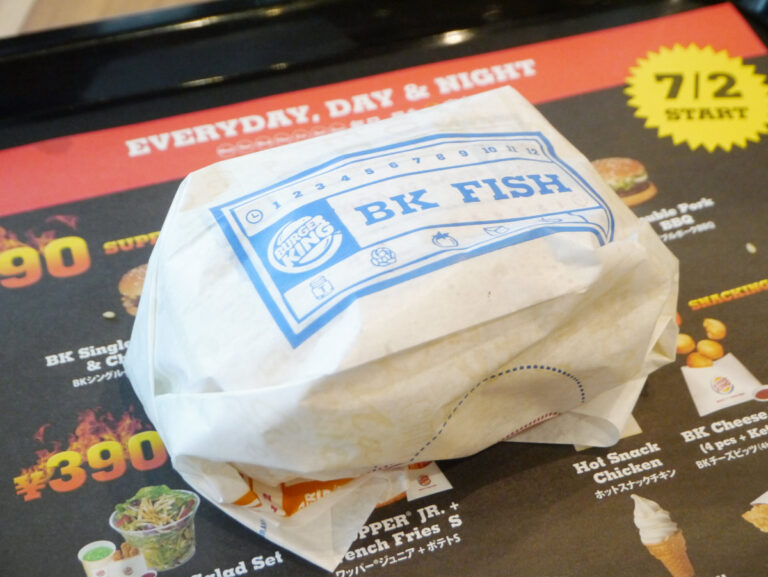 The image shows the burgerking fish sandwich in the warp on the table and behind it there is burger king menu also.