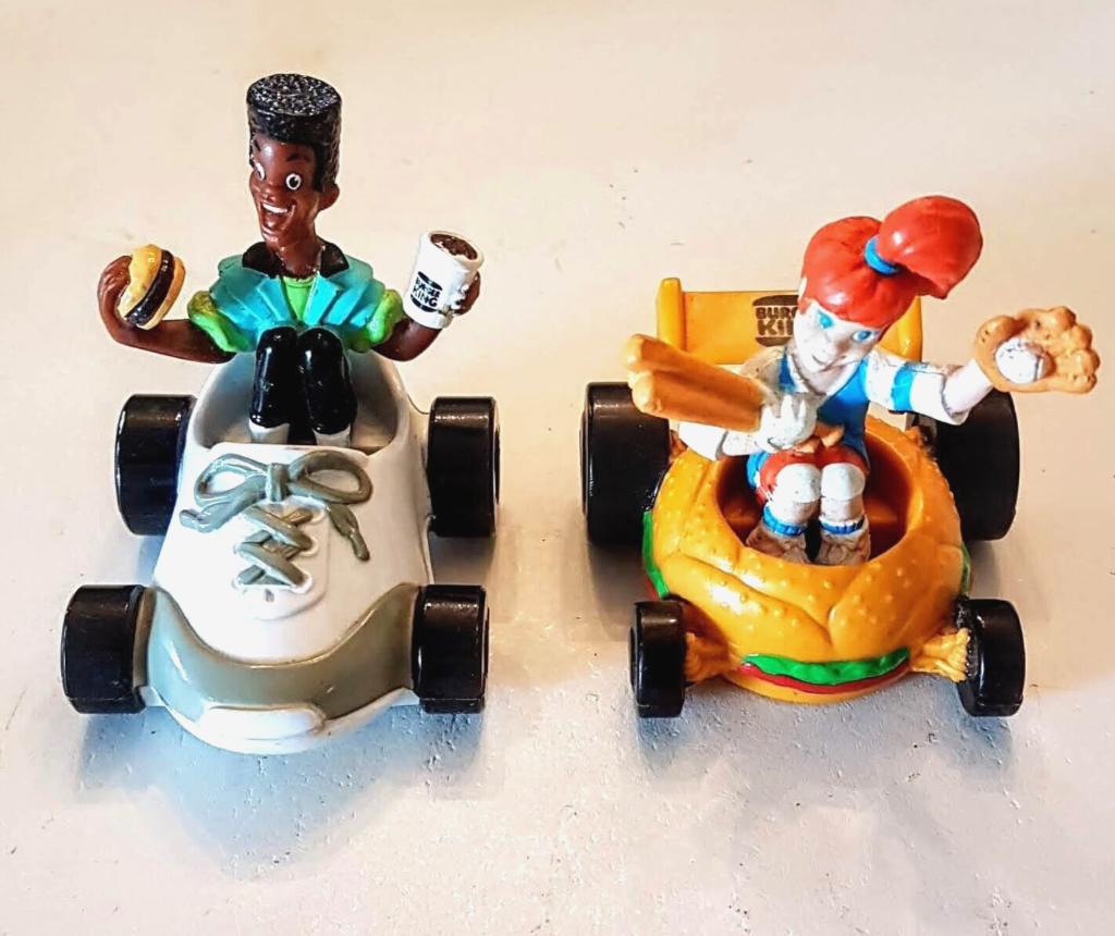 Two vintage Boomer
Burger King Kids Club toys. The first toy is a white race car with a black figure driving it. The figure is holding a hamburger and a coffee cup. The second toy is a yellow race car shaped like a hamburger bun. The figure inside is holding a baseball bat and a baseball glove.